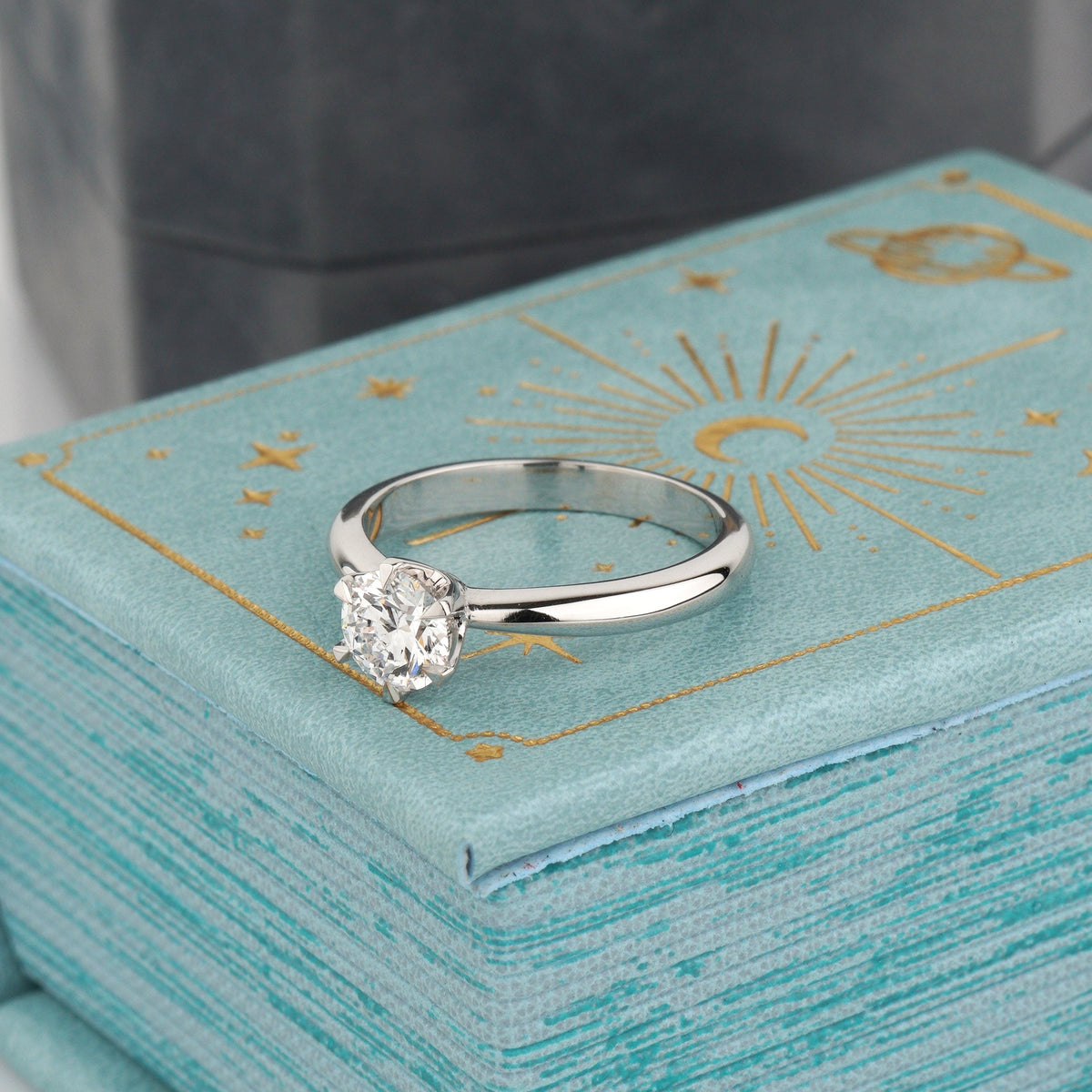 A white gold engagement ring with a round-cut clear gemstone rests on a light blue textured box with gold celestial illustrations-solitaire engagement ring-igi diamond engagement ring -classic engagement ring-round solitaire ring