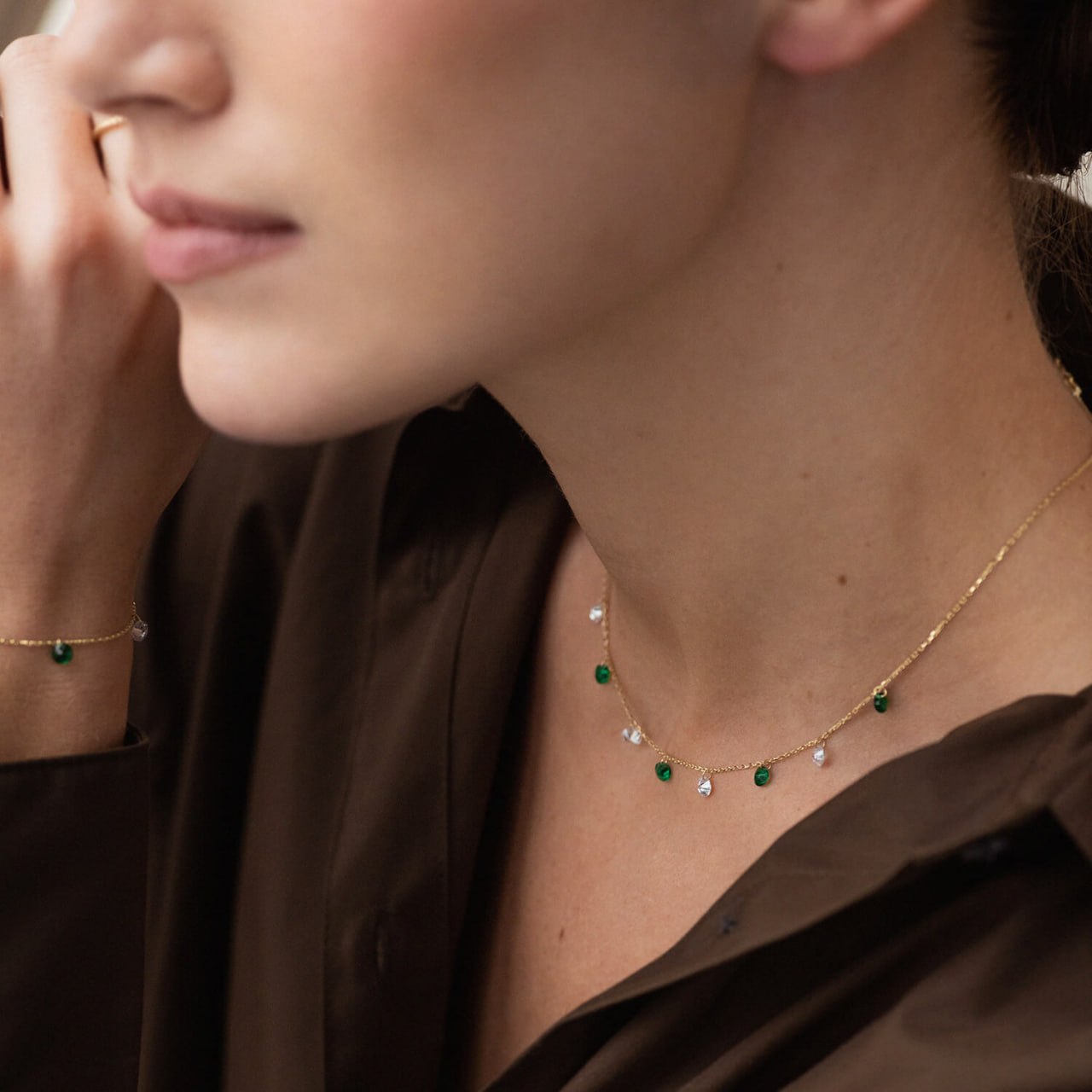 A close-up lifestyle shot of a woman wearing a delicate gold necklace with small green and clear gemstone accents, along with a matching gold bracelet on her wrist