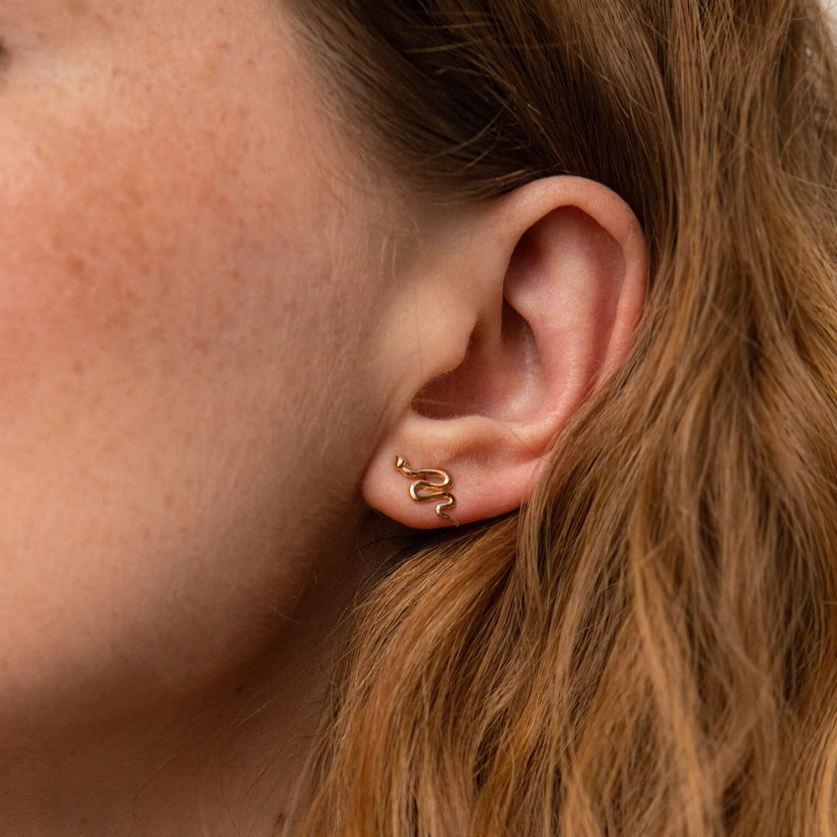 A close-up of a person's ear wearing a small gold snake-shaped stud earring-Metal: 14k Yellow ot Rose Gold (585)
Stone: None
Size: 1.3 x 0.7 cm