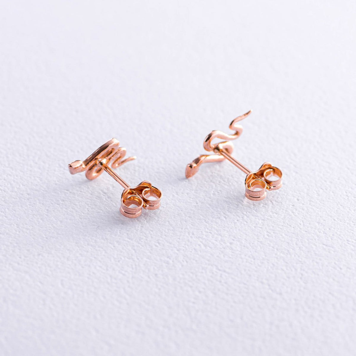 A pair of rose gold stud earrings with a unique coiled snake design, displayed on a textured white surface-snake earrings
gold stud earrings
14k gold earrings