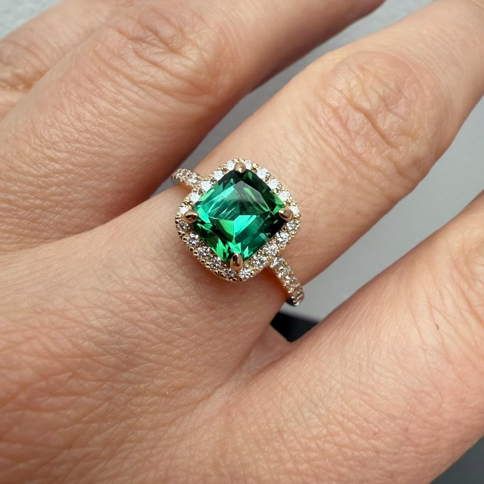 A cushion-cut emerald green gemstone ring with a halo of small clear stones and a diamond-accented band, shown worn on a hand-Metal: 14k Yellow Gold (585), solid
Center Stone: Natural Green Tourmaline, Cushion Cut, 1.7 ct
Accent Stones: Natural Diamonds, VVS2/F, 0.41 ct total
Setting: Diamond halo with pavé band
