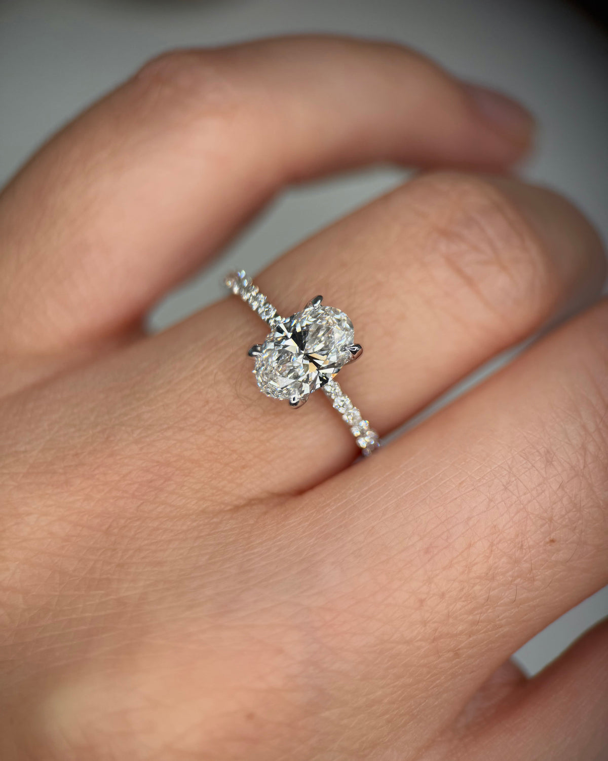 A close-up of a hand wearing a silver-toned ring with a large round-cut center stone and smaller stones along the band, set against a soft gray background-solid gold engagement ring
diamond proposal ring