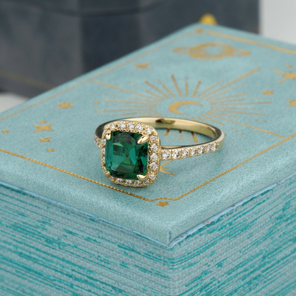 A yellow gold ring featuring a square-cut green emerald center stone surrounded by a halo of small clear pavé stones-statement engagement ring-custom engagement ring-green stone engagement ring