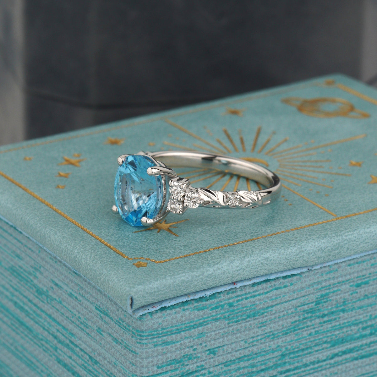 A silver ring with a light blue oval gemstone and small clear accent stones, displayed on a textured turquoise box with gold celestial line art-nature inspired ring-colored gemstone ring-topaz gold ring