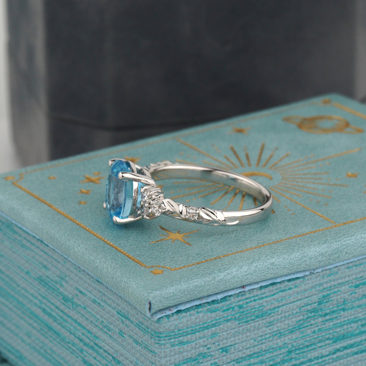 A white gold ring with a rectangular light blue gemstone and small side stones, displayed on a turquoise box with gold celestial designs-Center Stone: Natural Swiss Blue Topaz, Oval Cut, 1.5 ct