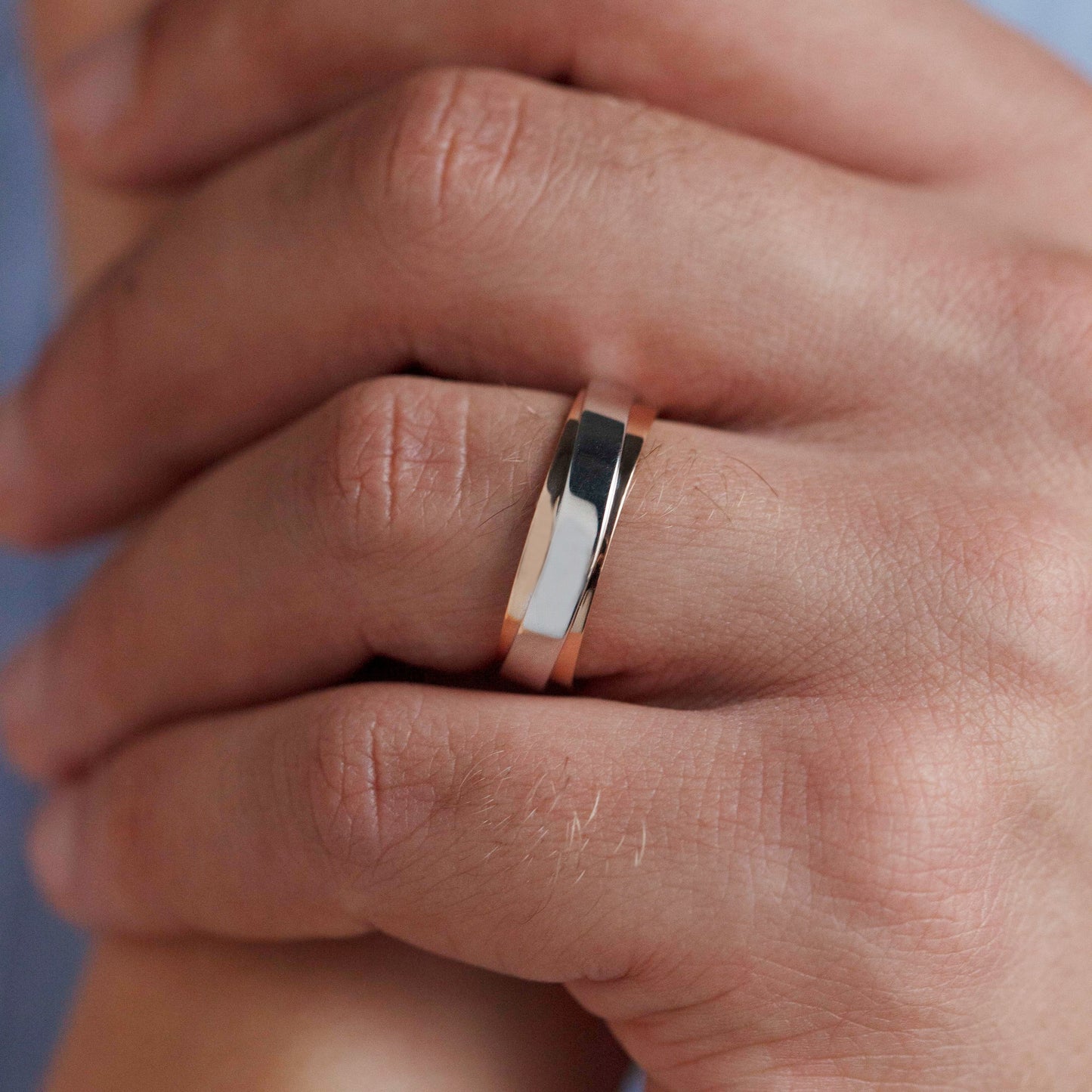 A close-up of a person's hands resting together, with a slim metallic band ring worn on one finger. The ring has a polished silver-toned center with a rose gold-toned outer edge-mens wedding band gold -unique wedding band